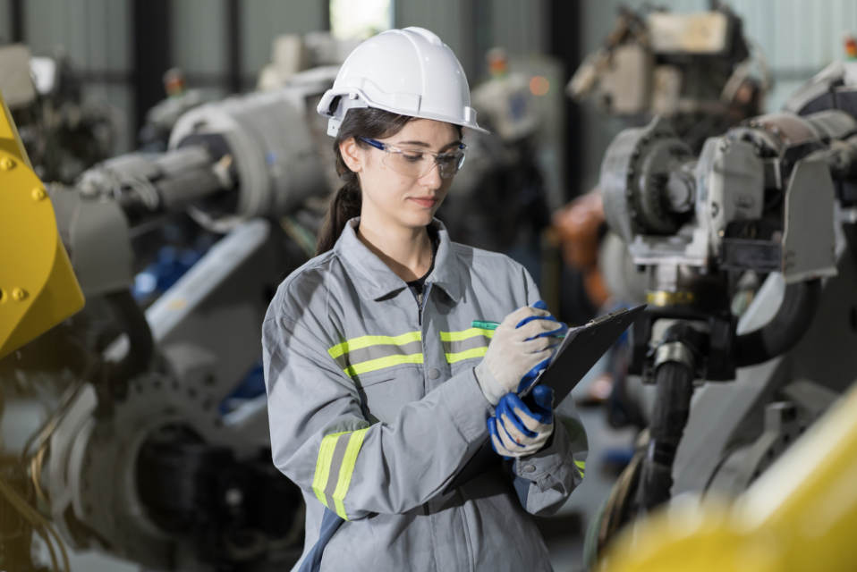 A woman in protextive gear records NDT data on a clipboard in front of machinery