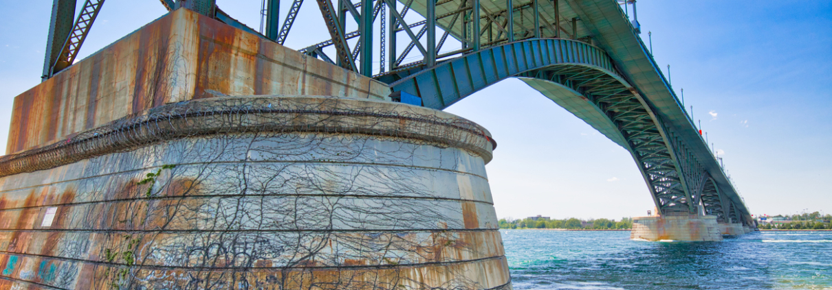 Steel bridge structure extending over a river with a large concrete support pier covered in vegetation and visible signs of weathering.