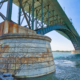 Steel bridge structure extending over a river with a large concrete support pier covered in vegetation and visible signs of weathering.