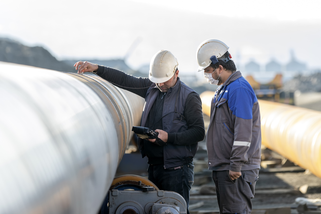 Two industrial technicians wearing hard hats inspect a large pipeline while reviewing equipment data at an outdoor work site.